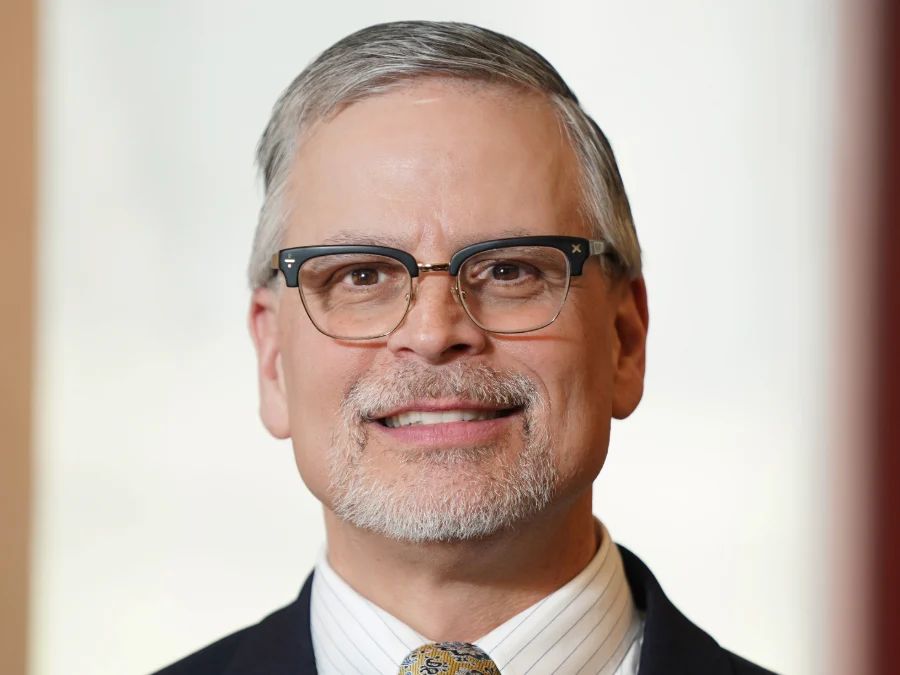 A middle-aged man with gray hair and a goatee wearing glasses, a suit, a striped shirt, and a patterned tie, smiling at the camera against a blurred background.