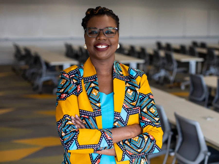 A woman wearing glasses and a bright yellow patterned blazer stands with arms crossed in a conference room with rows of empty chairs and tables.