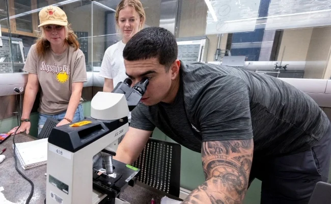 A man looks into a microscope while two women observe him from behind in a laboratory setting.