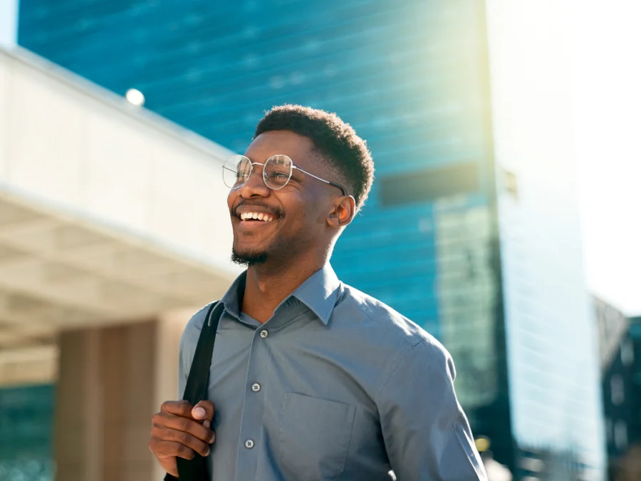 A man wearing glasses and a gray shirt smiles while standing outdoors in a city, holding a shoulder bag, with modern buildings in the background.