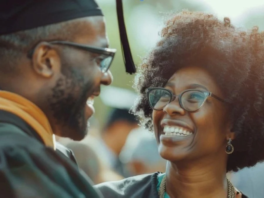 Two people wearing graduation caps and gowns smile at each other outdoors during a graduation ceremony.