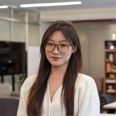 A woman with long brown hair and glasses, wearing a white top, stands indoors in an office setting with bookshelves and modern furniture in the background.