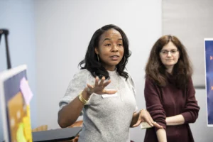 Two women stand indoors; one gestures as she speaks, while the other listens attentively. Posters and presentation materials are visible in the background.