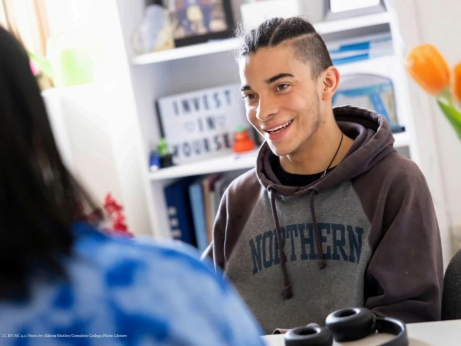 A young person in a “Northern” hoodie smiles while talking to someone across a desk; a sign reading “Invest in Yourself” is visible in the background.